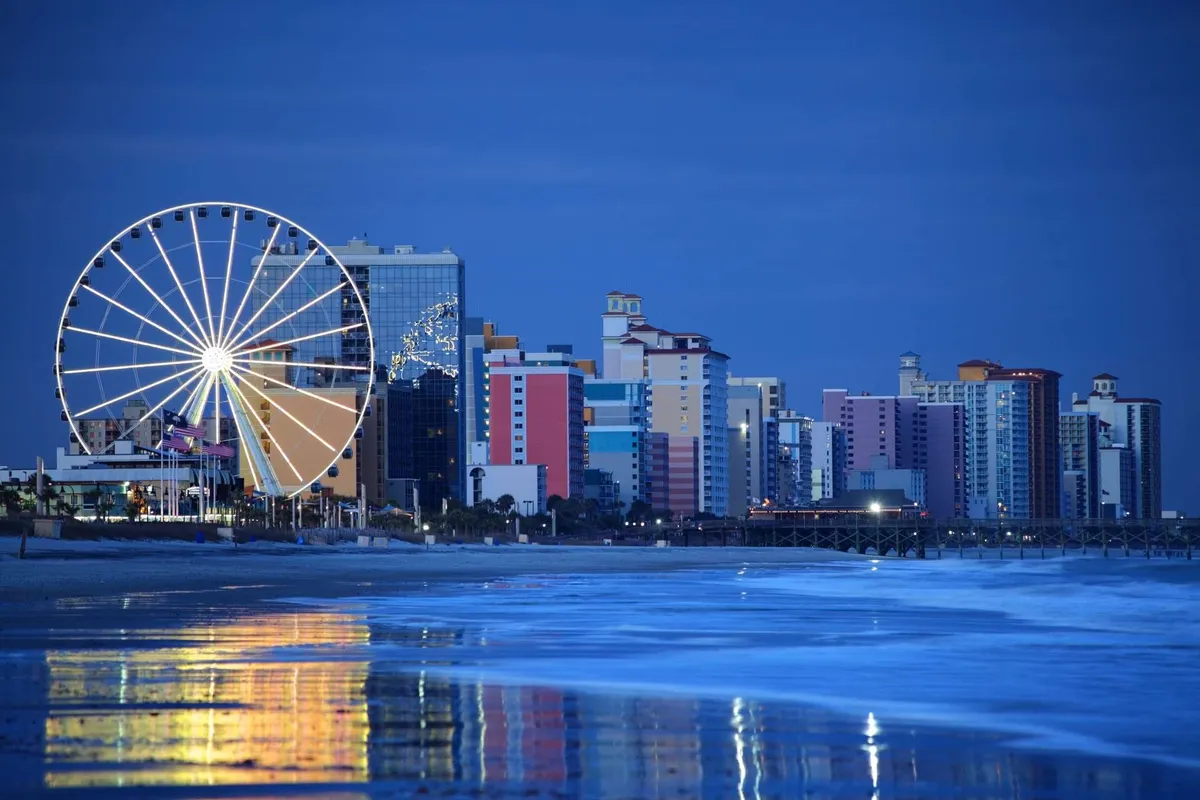A vibrant scene featuring the iconic Myrtle Beach Ferris wheel, colorful beachfront buildings, and the shimmering ocean at sunset. The reflection in the water and festive energy suggest the abundance of local attractions and lively family outings nearby.