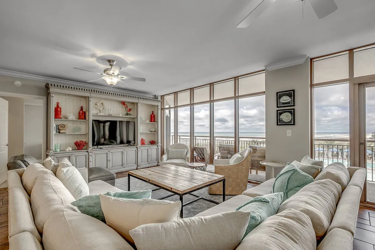 The image showcases a spacious, sunlit living area with a plush sectional sofa arranged for group gatherings, an entertainment center perfect for movie nights, expansive floor-to-ceiling windows revealing breathtaking balcony views, and inviting outdoor seating ideal for al fresco dining—all set against the backdrop of North Myrtle Beach.