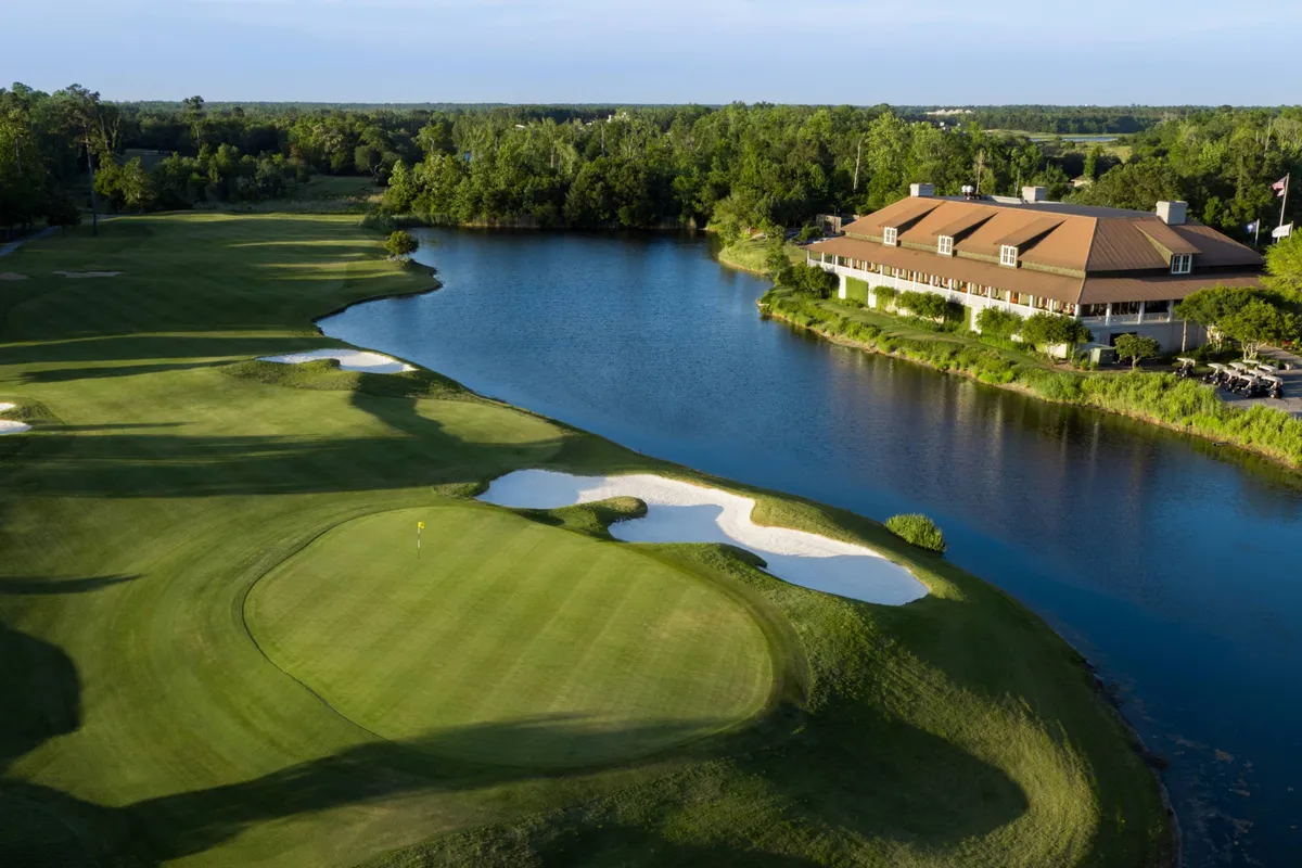 A sweeping view of the Azalea Sands Golf Club, showcasing an immaculately maintained course with lush greens, sand traps, and a scenic water feature. The clubhouse sits in the background and the sun shines over the landscape, inviting visitors out for a morning or afternoon round of golf.