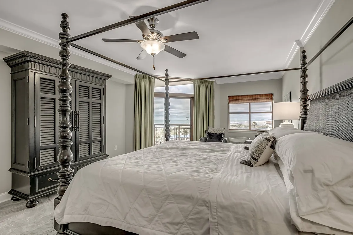 A restful bedroom with a four-poster bed, black armoire for storage, large windows with flowing curtains, and a ceiling fan. The room is bright and airy, providing a tranquil retreat with ample storage solutions.