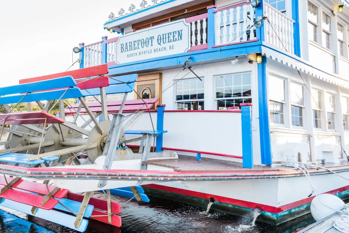 A festive exterior shot featuring a colorful paddlewheel riverboat docked by the shore. The scene highlights opportunities for dining, water tours, and live music, with lively signage and a festive façade that hint at the vibrant local nightlife and entertainment nearby.