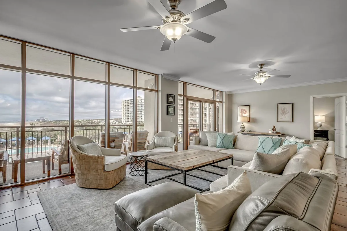 A spacious, inviting living area with a comfortable seating arrangement positioned around a wooden coffee table. Floor-to-ceiling windows frame a stunning ocean view, bathing the room in natural light. The modern, coastal-inspired furnishings echo the serene seaside setting.