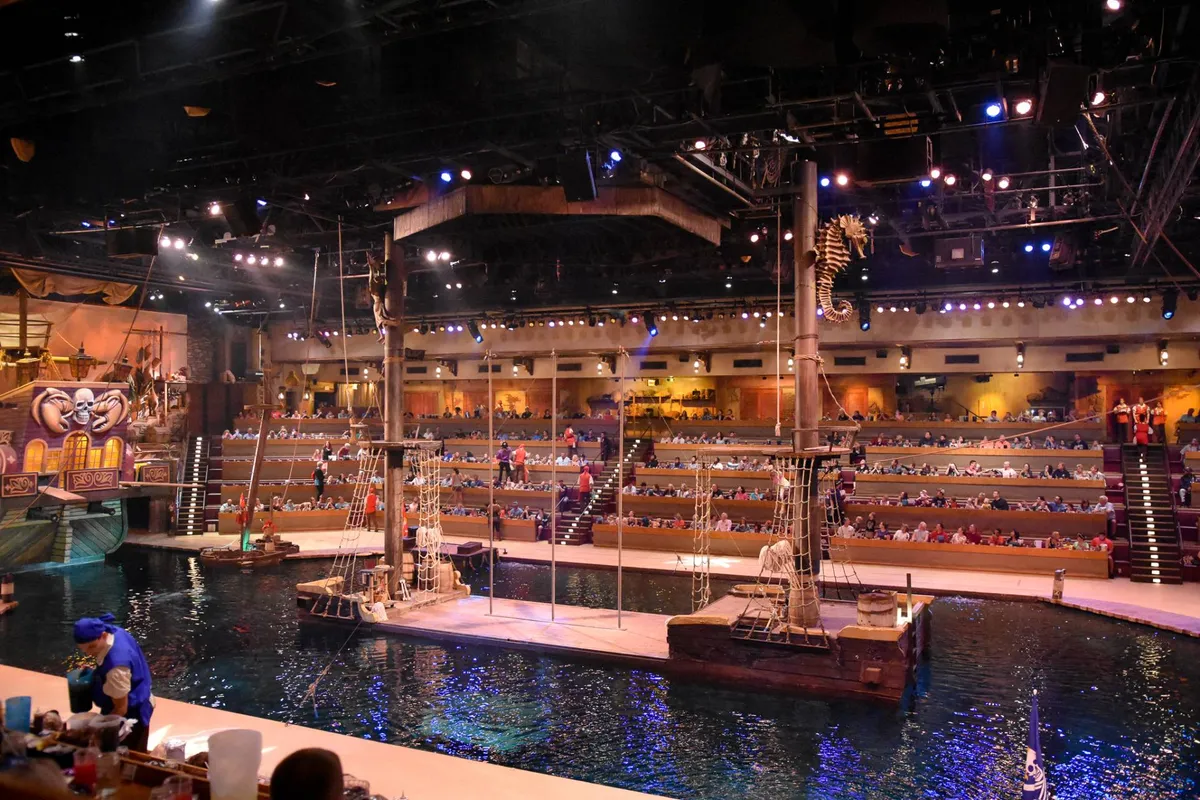 This interior photograph captures the lively atmosphere of a themed dinner show, featuring a large pirate ship, audience seating, and a dramatic water performance stage. The scene is filled with anticipation and immersive storytelling.