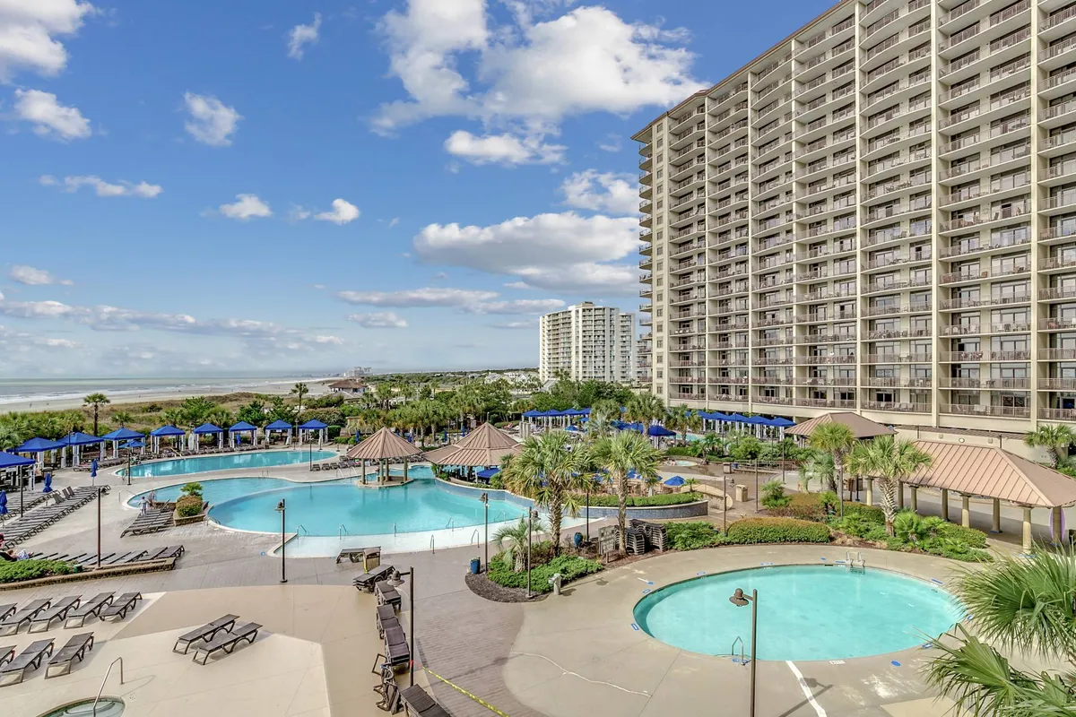 A broad, sunlit view of the resort’s main pool complex, complete with winding pools, palm trees, lounge chairs, and cabanas. The architecture and landscaping showcase the size and variety of the outdoor amenities available to guests.