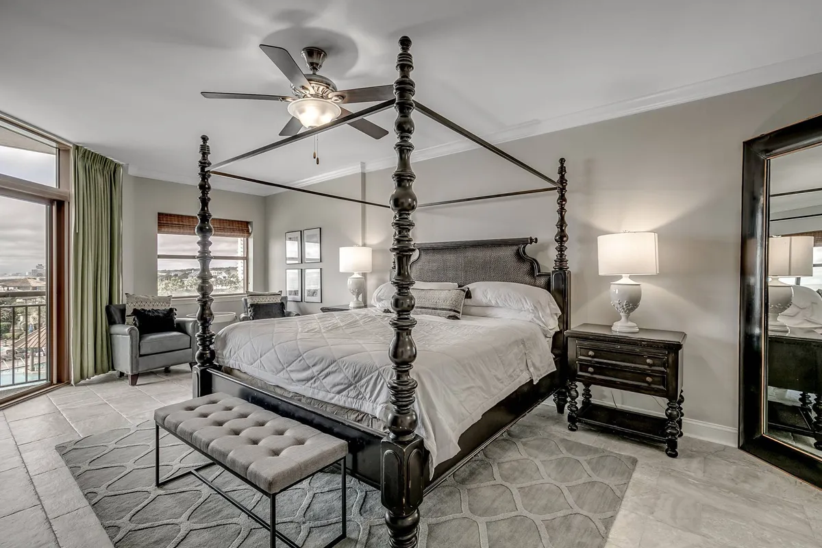 A serene bedroom featuring a four-poster bed, a cozy sitting area, large windows that let in natural light, and a decorative mirror. The space exudes tranquility with soft tones and plush furnishings.