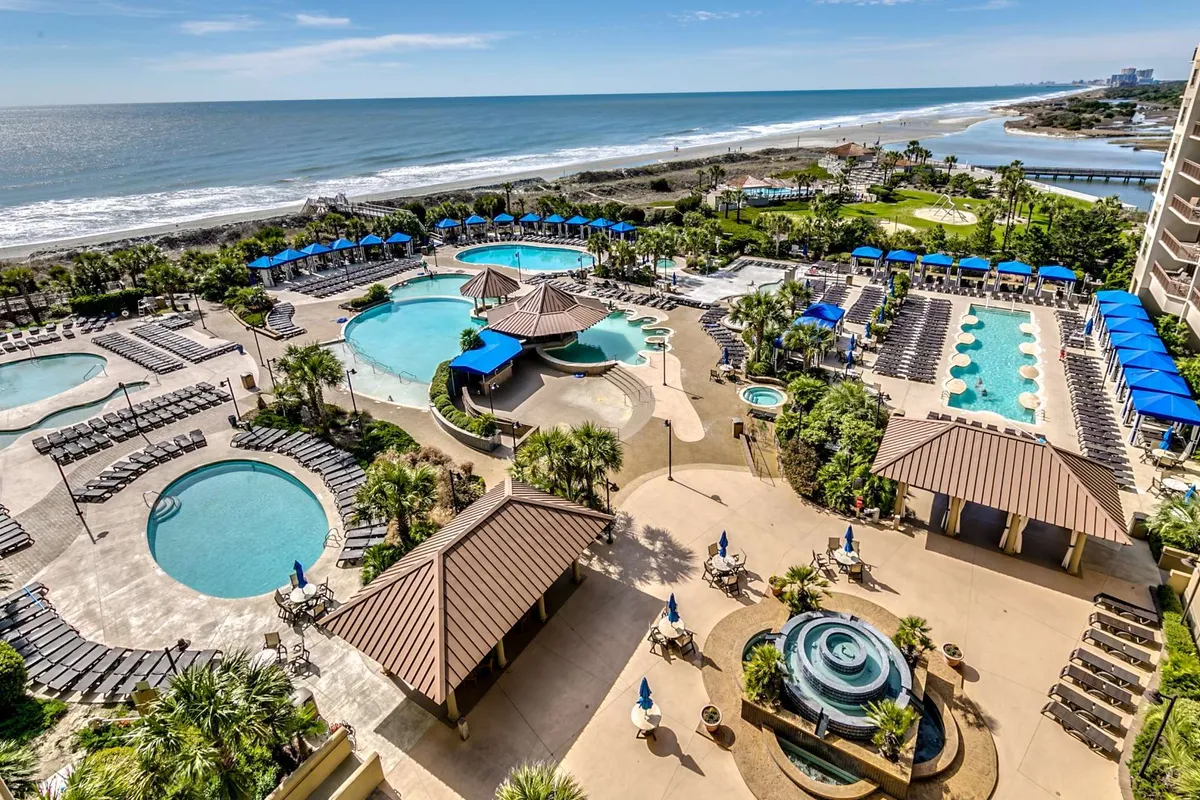 A vibrant exterior shot of a sprawling beachfront pool complex, dotted with palm trees and inviting lounge areas. Guests can be seen relaxing by the water, and the resort’s proximity to the ocean is evident. The atmosphere evokes vacation luxury, perfect for both day and evening enjoyment.