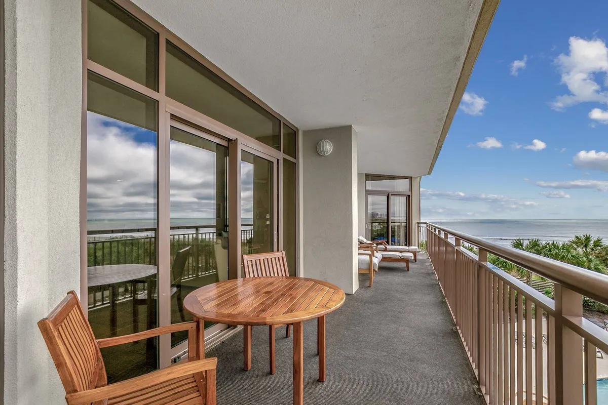 A serene covered balcony furnished with a wooden table and chairs overlooks an expansive ocean view. Palm trees and the gentle ocean horizon set a relaxed, inviting scene for outdoor dining or sunset relaxation.