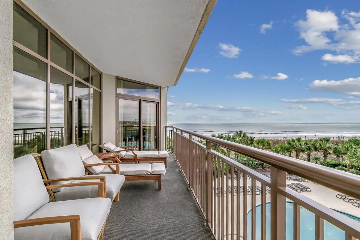 This image captures a relaxing balcony scene with comfortable seating, offering a sweeping view of both the ocean and the resort's expansive pool area below. The tranquil setting merges the vibrant pool complex with the allure of the beach just beyond.