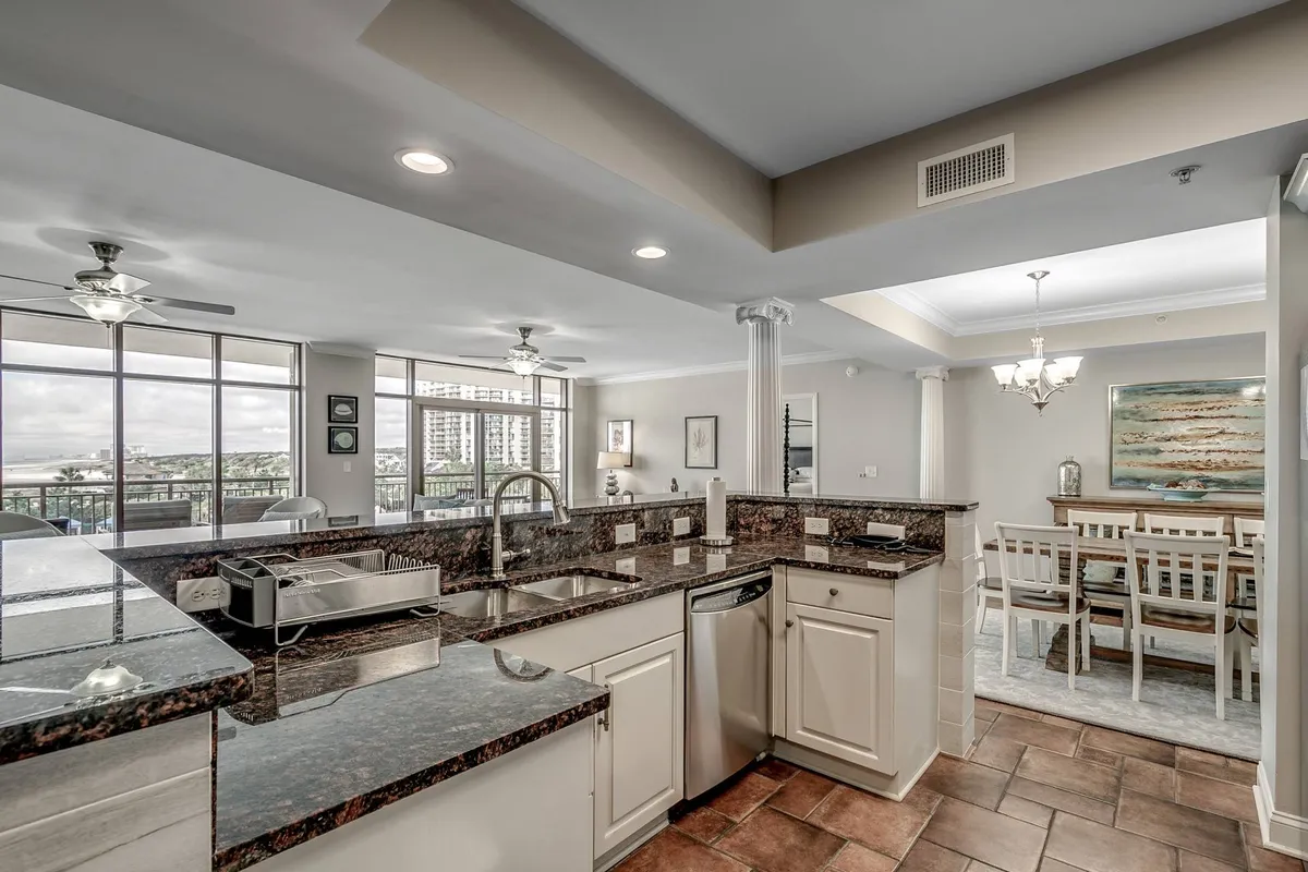 An open kitchen seamlessly flows into a modern dining area. Granite countertops, a kitchen island with seating, and ample lighting create a welcoming space for group cooking and socializing. The overall palette and design reinforce the coastal theme.