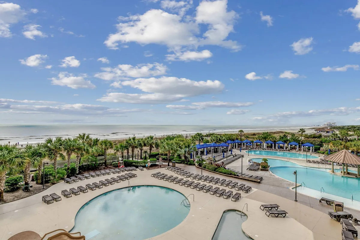 An exterior view of swimming pools surrounded by lounge chairs, palm trees, and blue canopies, with the beach visible in the background. The atmosphere is vibrant and resort-like, inviting guests to enjoy poolside or beach amenities at any time.